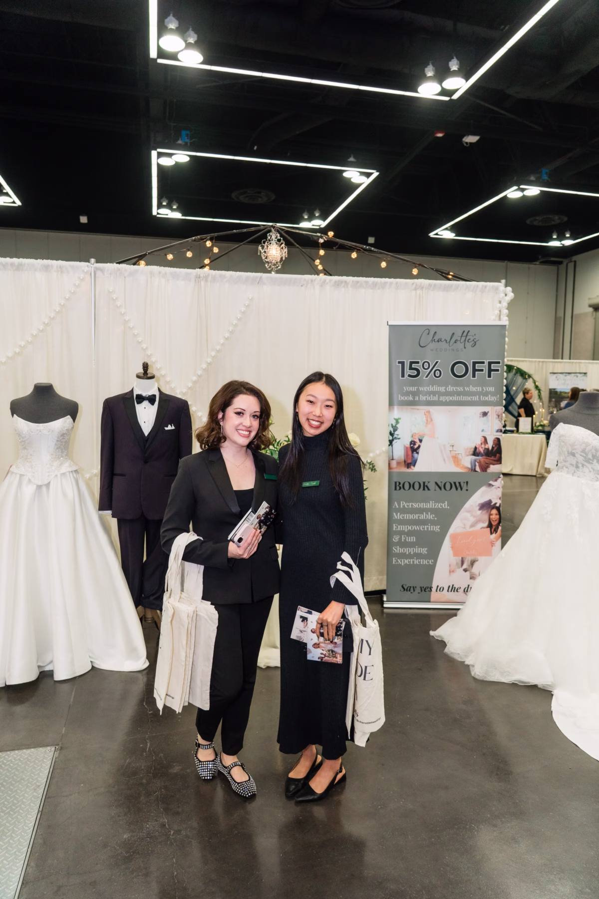Two women pose at Charlottes bridal boutique booth featuring wedding dresses and 15% off promotion at Portland Wedding Show 2026