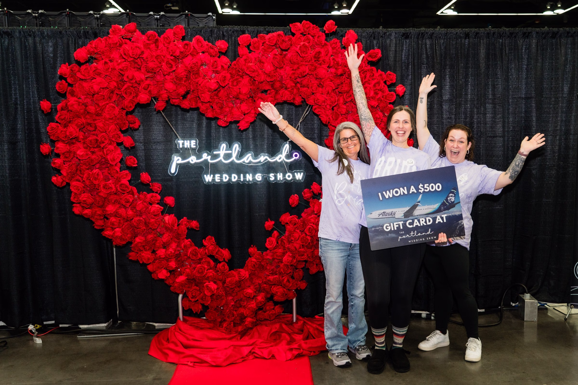 Three excited women celebrate winning a $500 Alaska Airlines gift card at The Portland Wedding Show 2026 in front of a large red heart-shaped floral backdrop