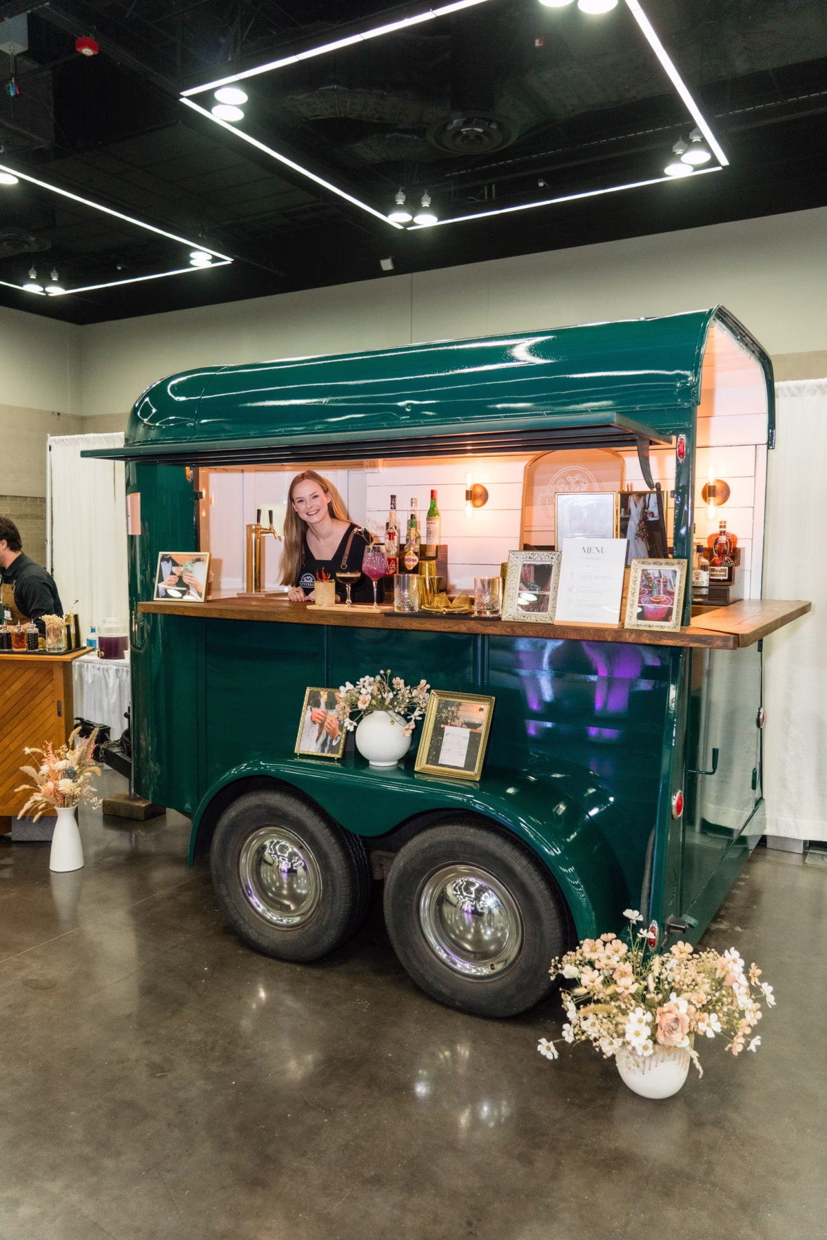 Green vintage mobile bar trailer with bartender at Portland Wedding Show 2026 bridal expo featuring elegant floral arrangements