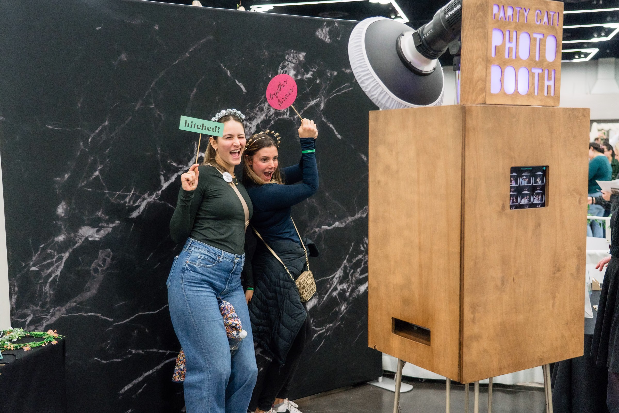 portland wedding show photo booth entertainment vendor - The Portland Wedding Show Two women pose with props at a wooden photo booth setup during the Portland Wedding Show bridal expo in Portland Oregon