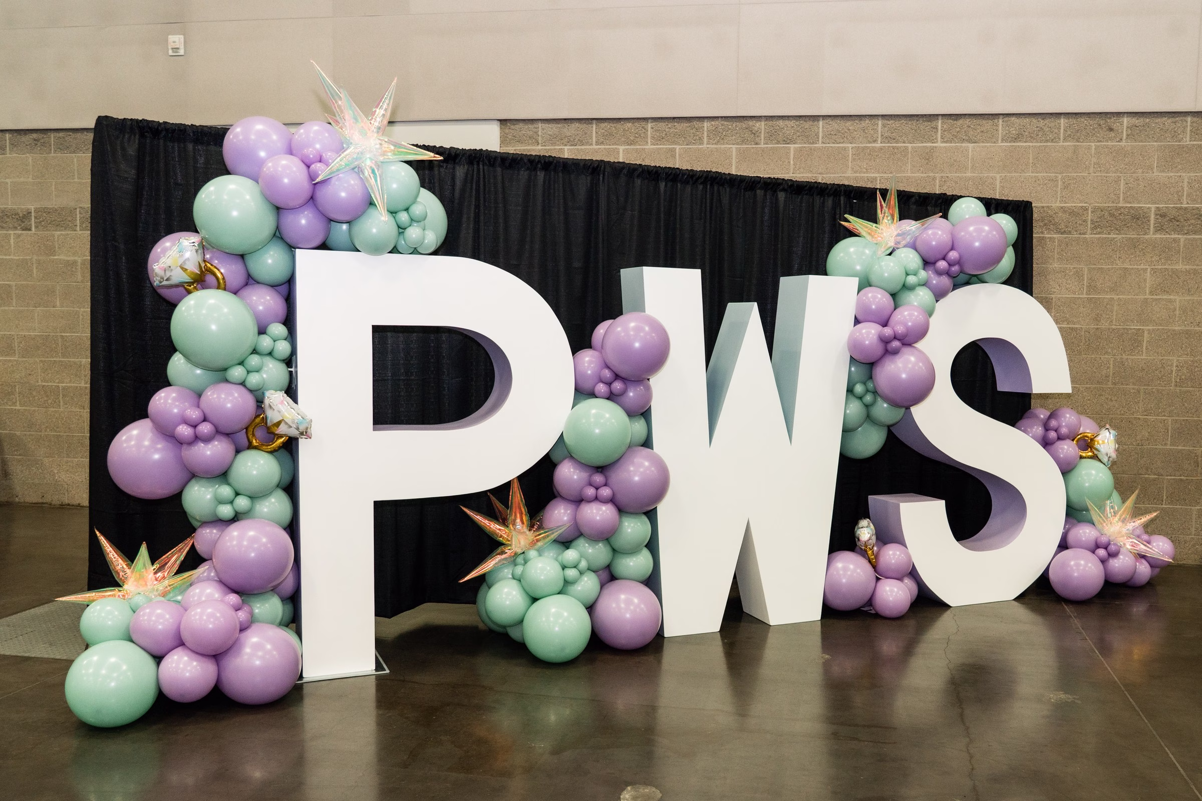 Large white PWS letters surrounded by purple and mint green balloon arrangements at Portland Wedding Show 2026 bridal expo